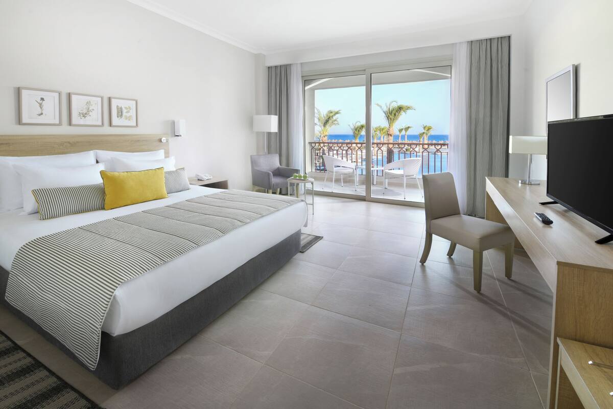 A hotel room with a bed, a desk with a TV, a chair, a balcony with a view of the sea, and artwork on the wall.