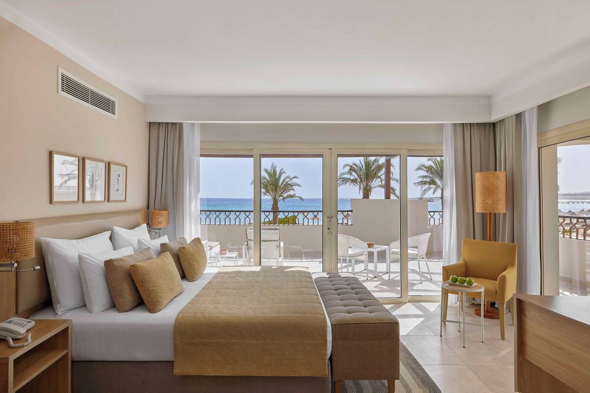 A hotel room with a bed, a desk, a chair, a balcony with a view of the sea, and a window with curtains.