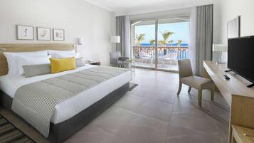 A hotel room with a large bed, a desk with a TV, a chair, a balcony with a view of the sea, and artwork on the wall.