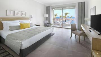 A hotel room with a bed, a desk with a TV, a chair, a balcony with a view of the sea, and artwork on the wall.