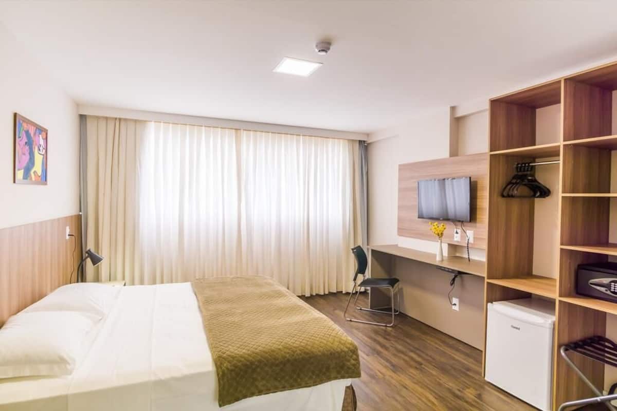 A hotel room with a bed, a desk, a chair, a TV, a wardrobe, and a small fridge.