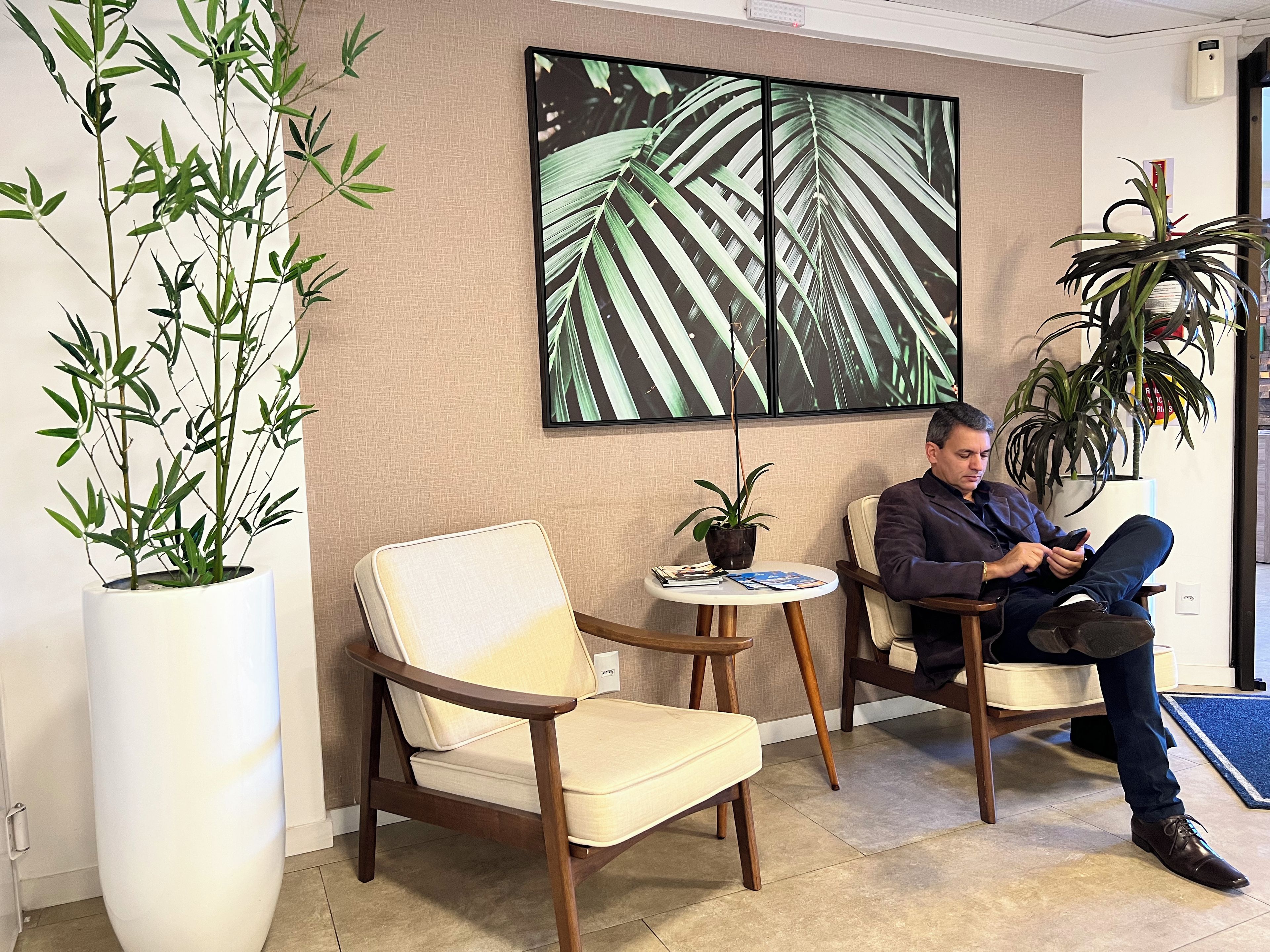 A man seated in a chair with a smartphone, a potted plant, and a framed artwork on the wall.