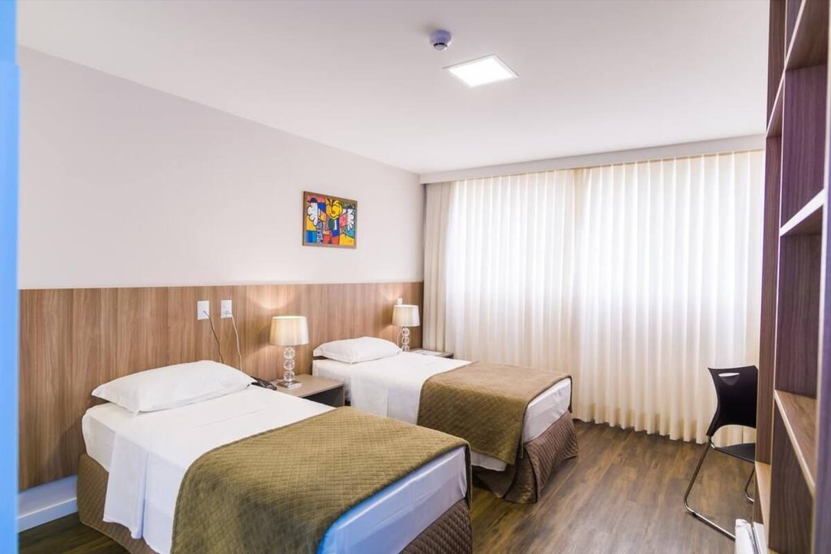 A hotel room with two beds, a desk, a chair, a window with curtains, and a ceiling light.