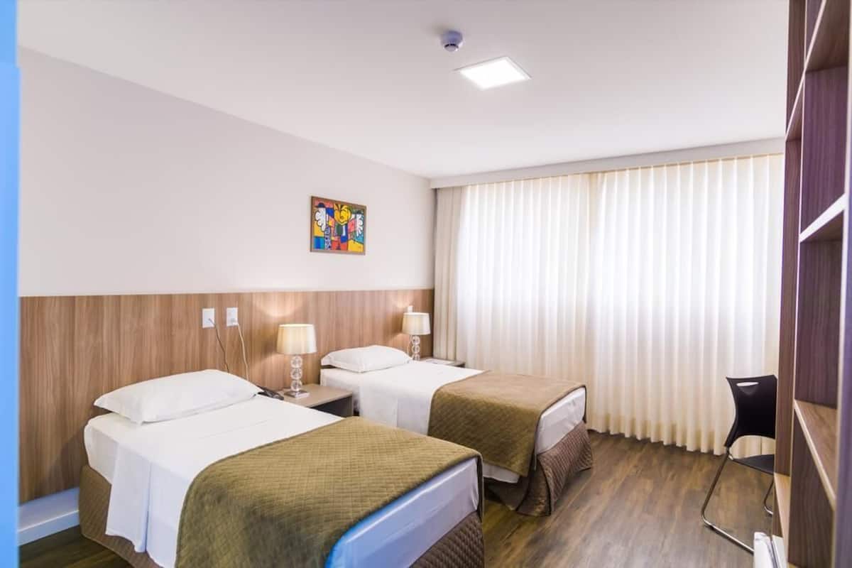 A hotel room with two beds, a desk, a chair, a window with curtains, and a ceiling light.