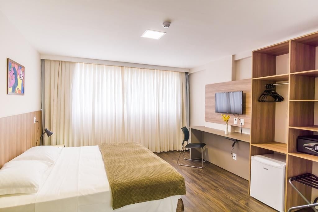 A hotel room with a bed, a desk, a chair, a TV, a wardrobe, and a small fridge.