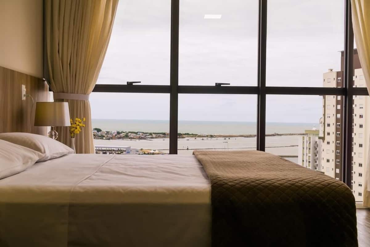A hotel room with a large window, a bed, and a view of a beach and buildings.