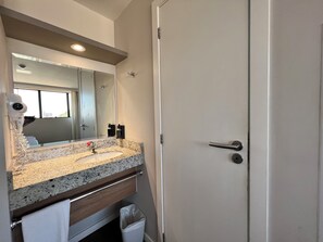 A bathroom with a granite countertop sink, a mirror, a towel rack, a trash bin, and a door with a handle. - Hotel 7 Itajai (Itajai)