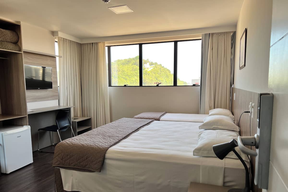 A hotel room with a bed, desk, chair, TV, and a window with a view of greenery.