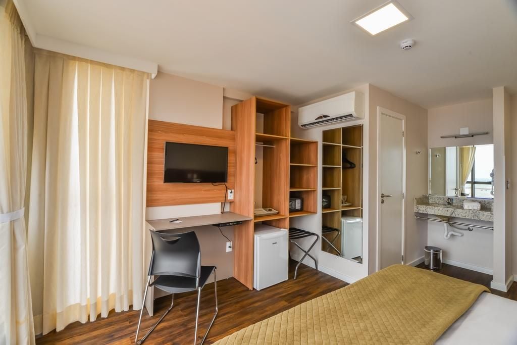 A bedroom with a bed, a desk, a chair, a TV, a wardrobe, and a bathroom with a mirror and sink.