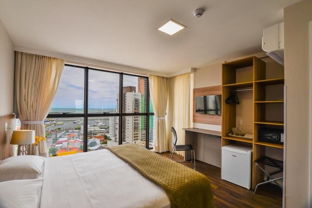 A hotel room with a bed, a desk, a chair, a TV, and a view of the city.