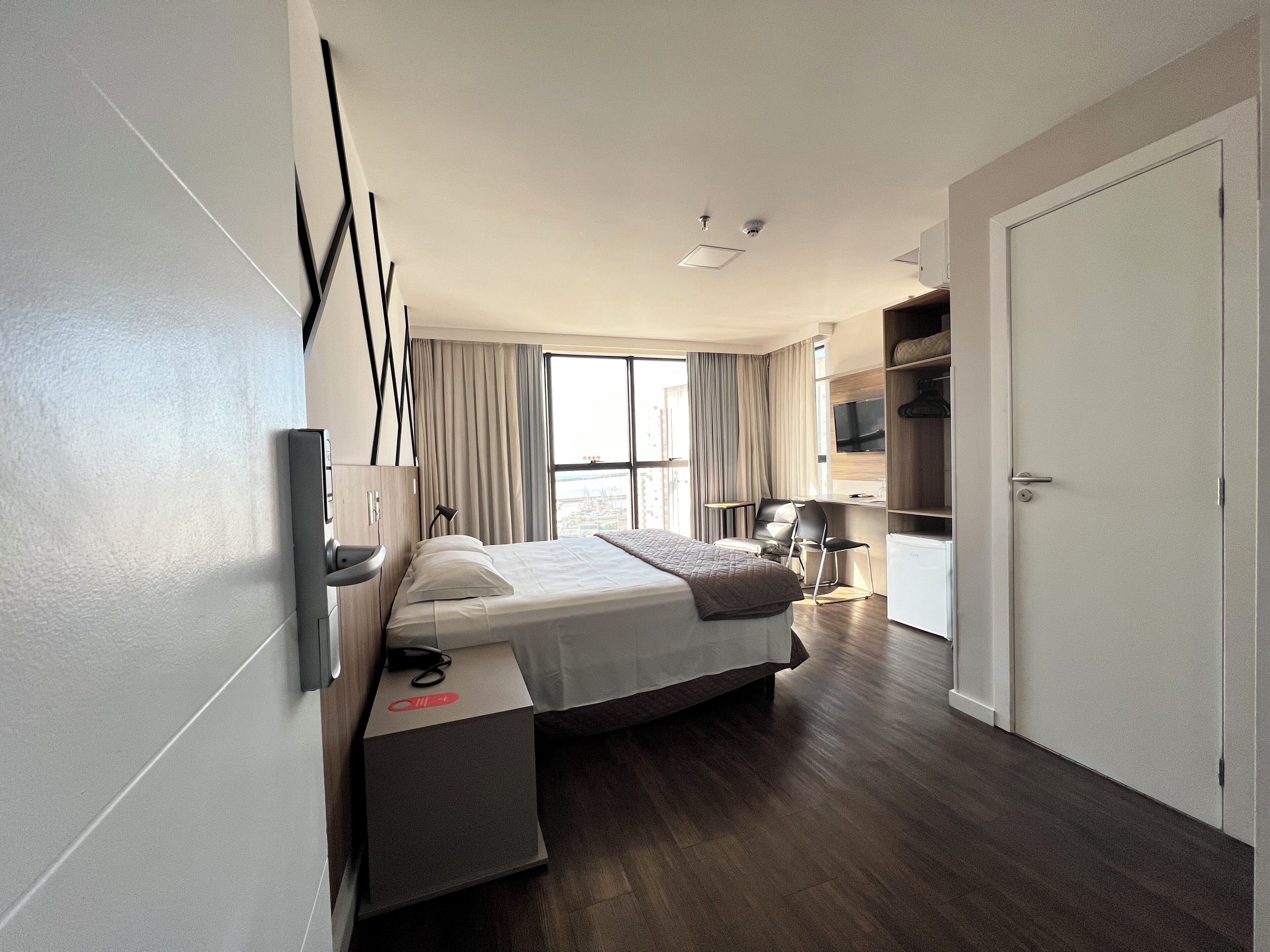 A modern hotel room with a bed, bedside table, TV, and a view of the city.