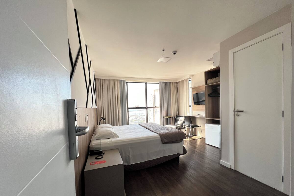 A modern hotel room with a bed, bedside table, TV, and a view of the city.