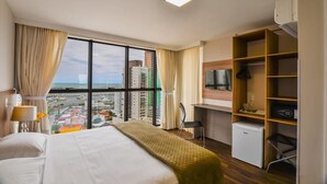 A hotel room with a bed, a desk, a chair, a TV, and a view of the city. - Hotel 7 Itajai (Itajai)