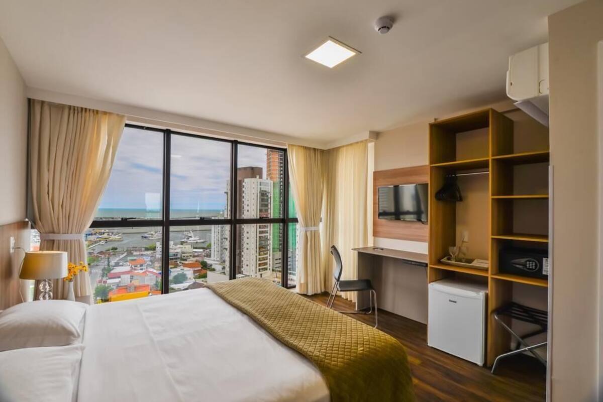 A hotel room with a bed, a desk, a chair, a TV, and a view of the city.