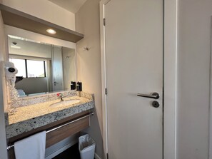 Une salle de bain avec un lavabo doté d’un plan de travail en granit, un miroir, un porte-serviettes, une poubelle et une porte avec une poignée.