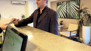 Un homme en costume au comptoir de la réception d’un hôtel, un écran d’ordinateur, une plante en pot et un tableau encadré accroché au mur.