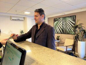 A man in a suit at a hotel reception desk, a computer monitor, a potted plant, and a framed picture on the wall. - Hotel 7 Itajai (Itajai)
