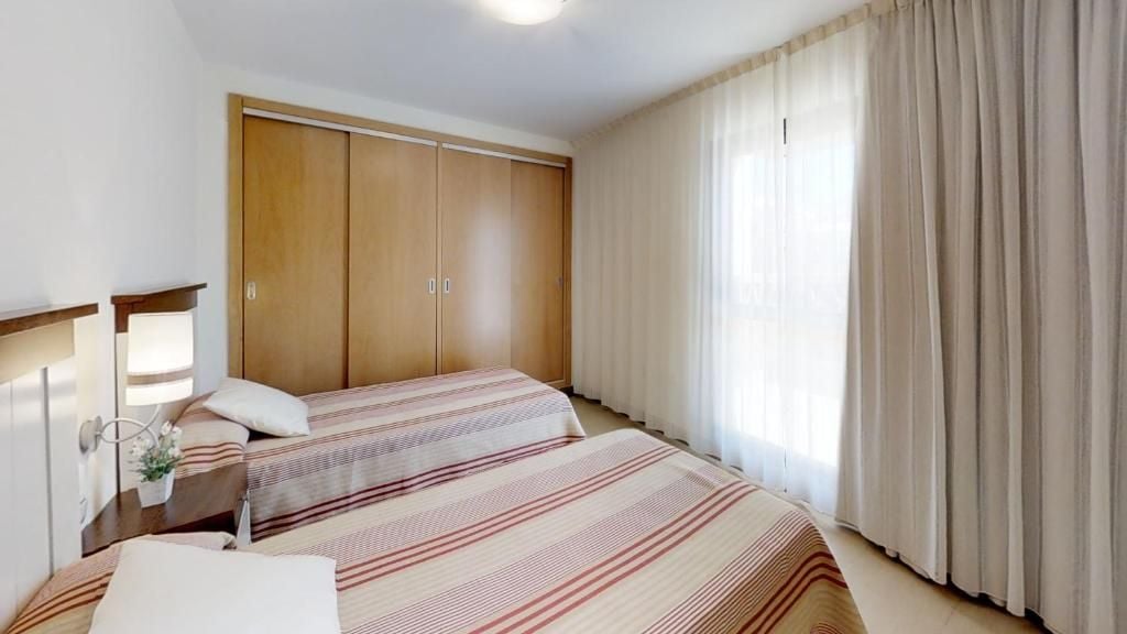 A bedroom with two beds, a wardrobe, a bedside table with a lamp, and a window with curtains.