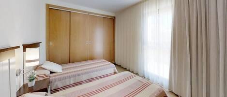 A bedroom with two beds, a wardrobe, a bedside table with a lamp, and a window with curtains.