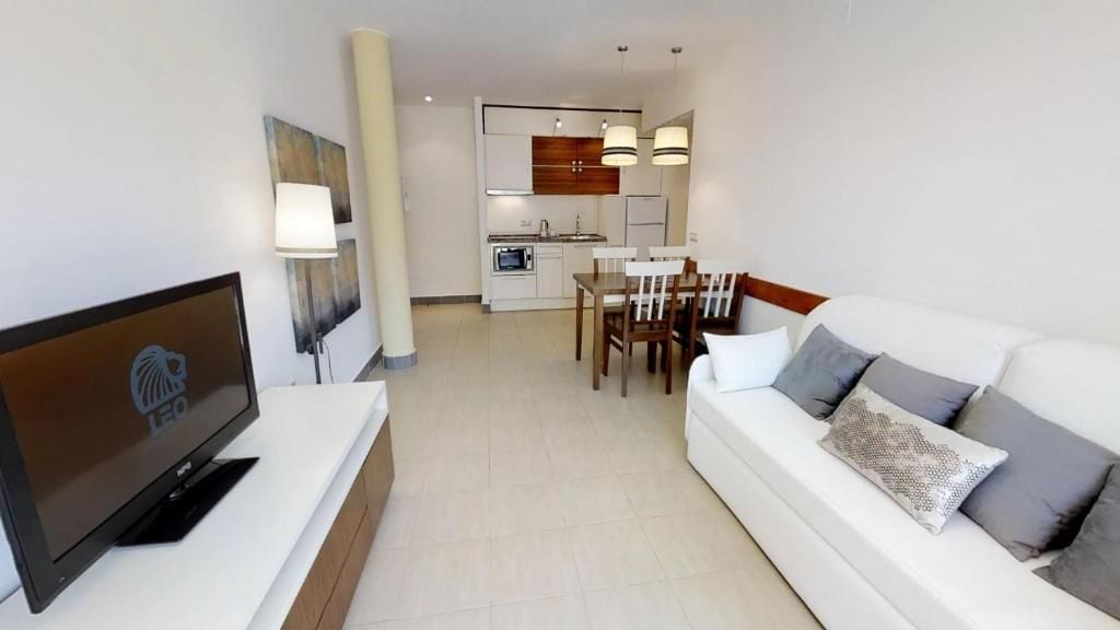 Una sala moderna con un televisor de pantalla plana, un sofá blanco con cojines estampados, un comedor con mesa y sillas, y una cocina al fondo.