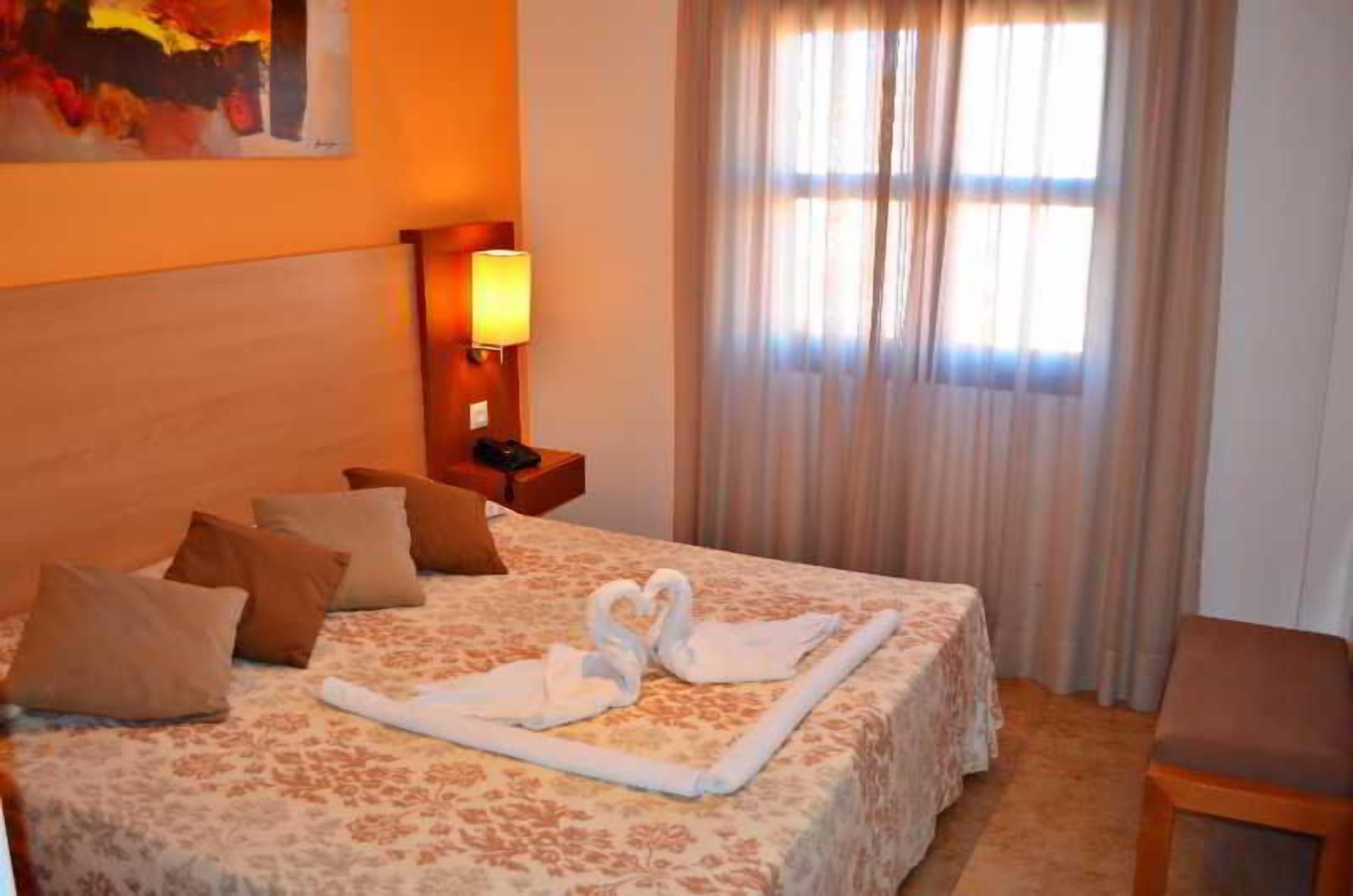 Habitación de hotel con cama, almohadas, lámpara de noche, una silla y una ventana con cortinas.