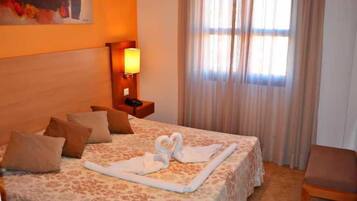 Habitación de hotel con cama, almohadas, lámpara de noche, una silla y una ventana con cortinas.
