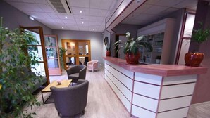 A reception area with a wooden desk, a grey sofa, and potted plants. - Hotel Les Bergeronnettes (Champagneux)