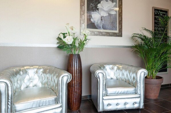 Un área de recepción con dos sillones tapizados plateados y acolchados, un alto jarrón marrón con flores blancas, una obra de arte floral enmarcada en la pared y una planta en maceta.