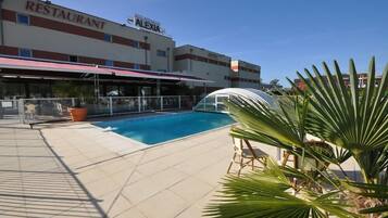 A swimming pool with a covered area, a restaurant named 'ALEXIA' in the background, and palm trees in the foreground.