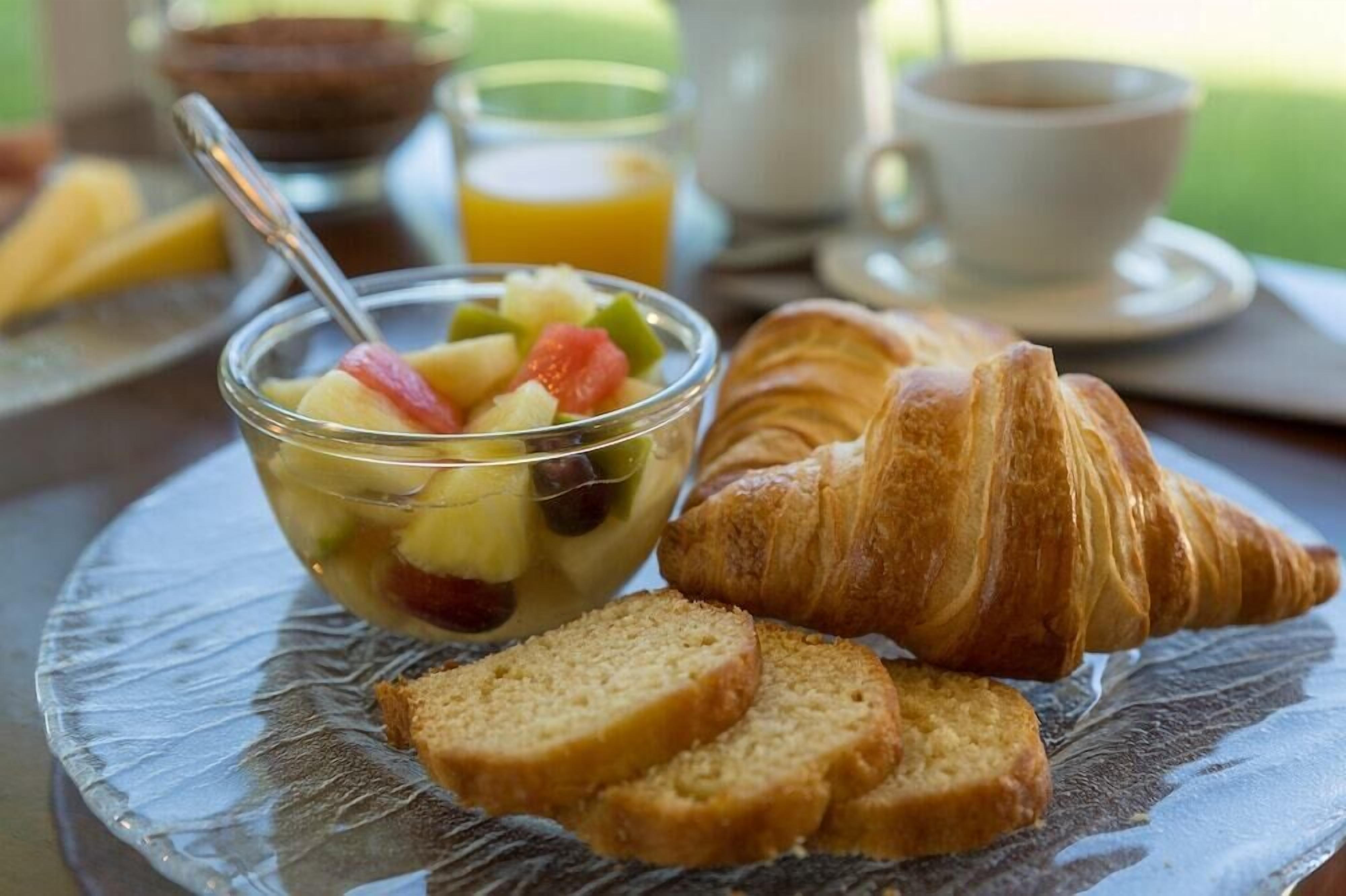 En morgenmadsopstilling med en skål frugtsalat, et croissanter og skiver af brød på en klar glasskål.