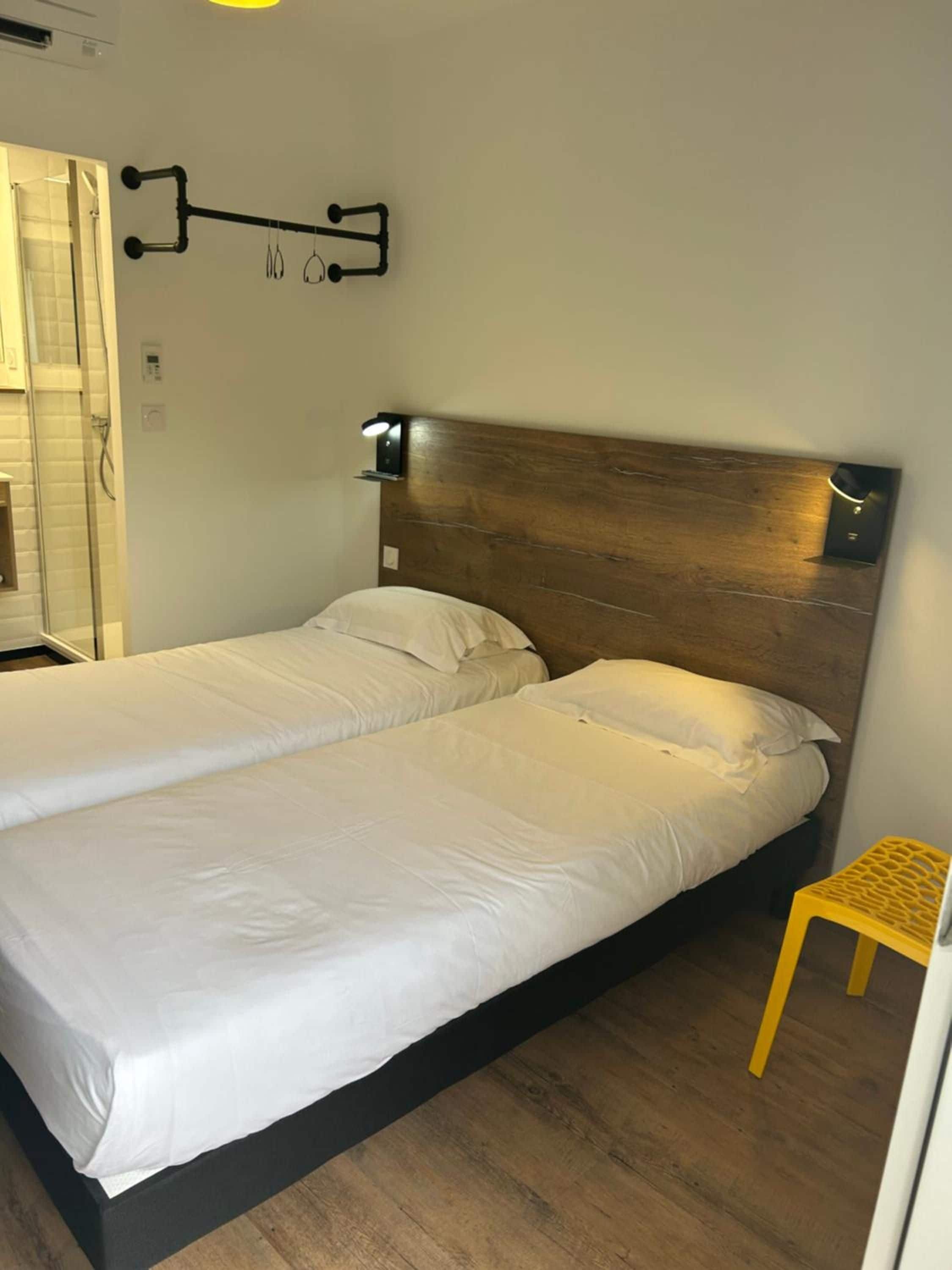 A double bed with a wooden headboard, a small yellow stool, and a wall-mounted towel rack.