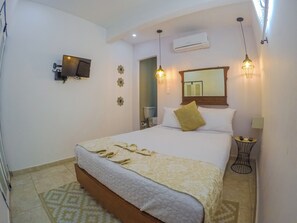 Una habitación de hotel con una cama, televisión, aire acondicionado y una mesita con lámpara.