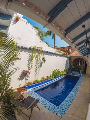 Un área de piscina con borde de baldosas, pared blanca con follaje y estructura de techo con azulejos.