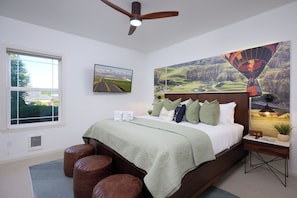 A bedroom with a bed, a ceiling fan, a window, a wall-mounted TV, and a large landscape painting.