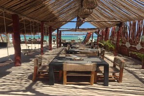 An outdoor dining area with thatched roof, wooden tables, and chairs, overlooking a beach. - Ensueno Holbox & Beach Club (Isla Holbox)
