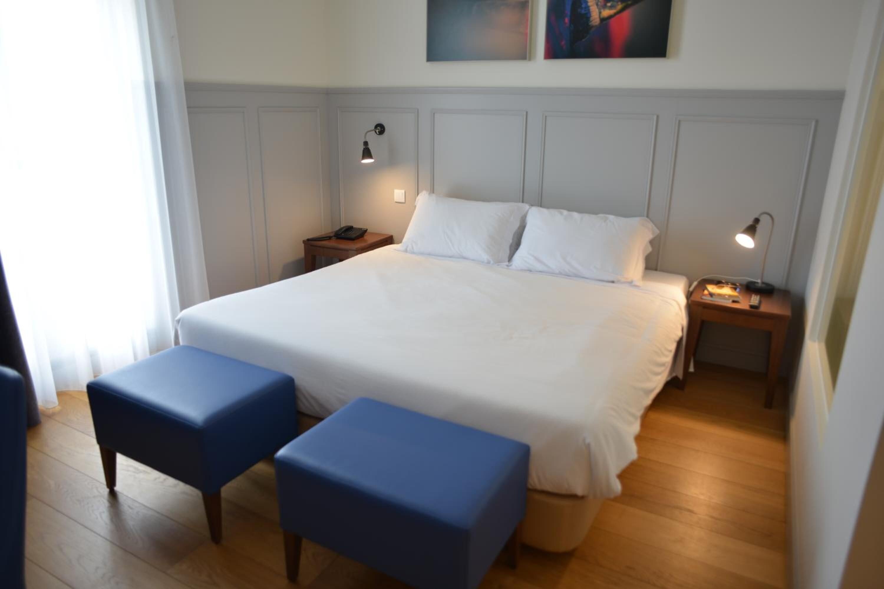 Habitación de hotel con una cama grande, dos mesitas de noche, un escritorio y dos otomanas azules.