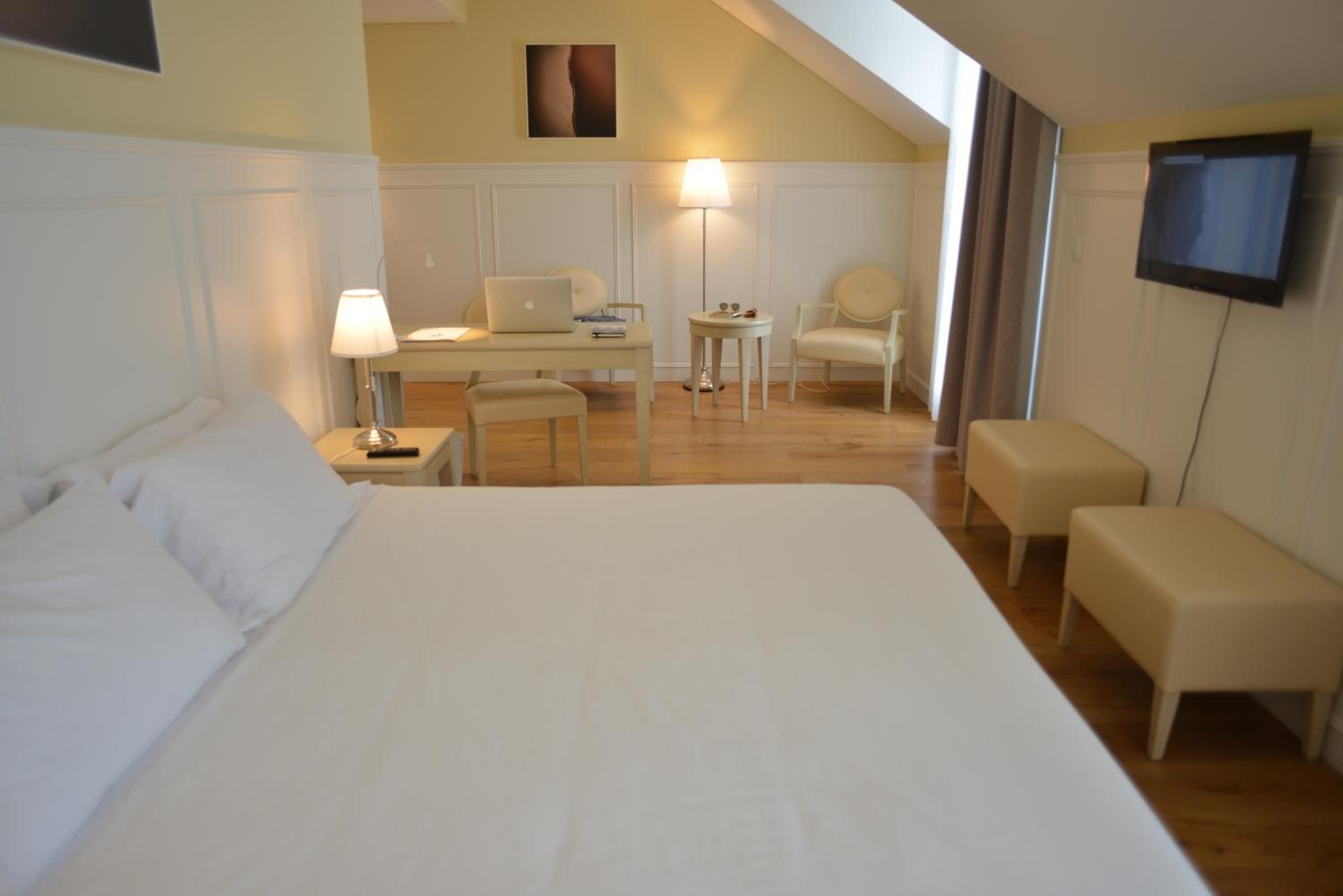 A hotel room with a large bed, a desk, a chair, a small table, a lamp, and a TV mounted on the wall.