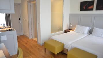 A hotel room with a bed, two yellow ottomans, a desk with a chair, a nightstand with a lamp, and a door leading to another room.
