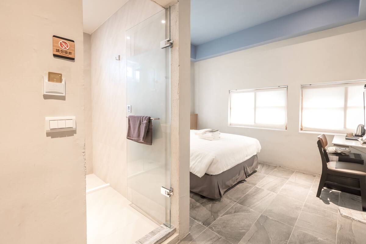 A modern bathroom with a glass shower enclosure, a bed with a white duvet, a desk with a chair, and a window with blinds.