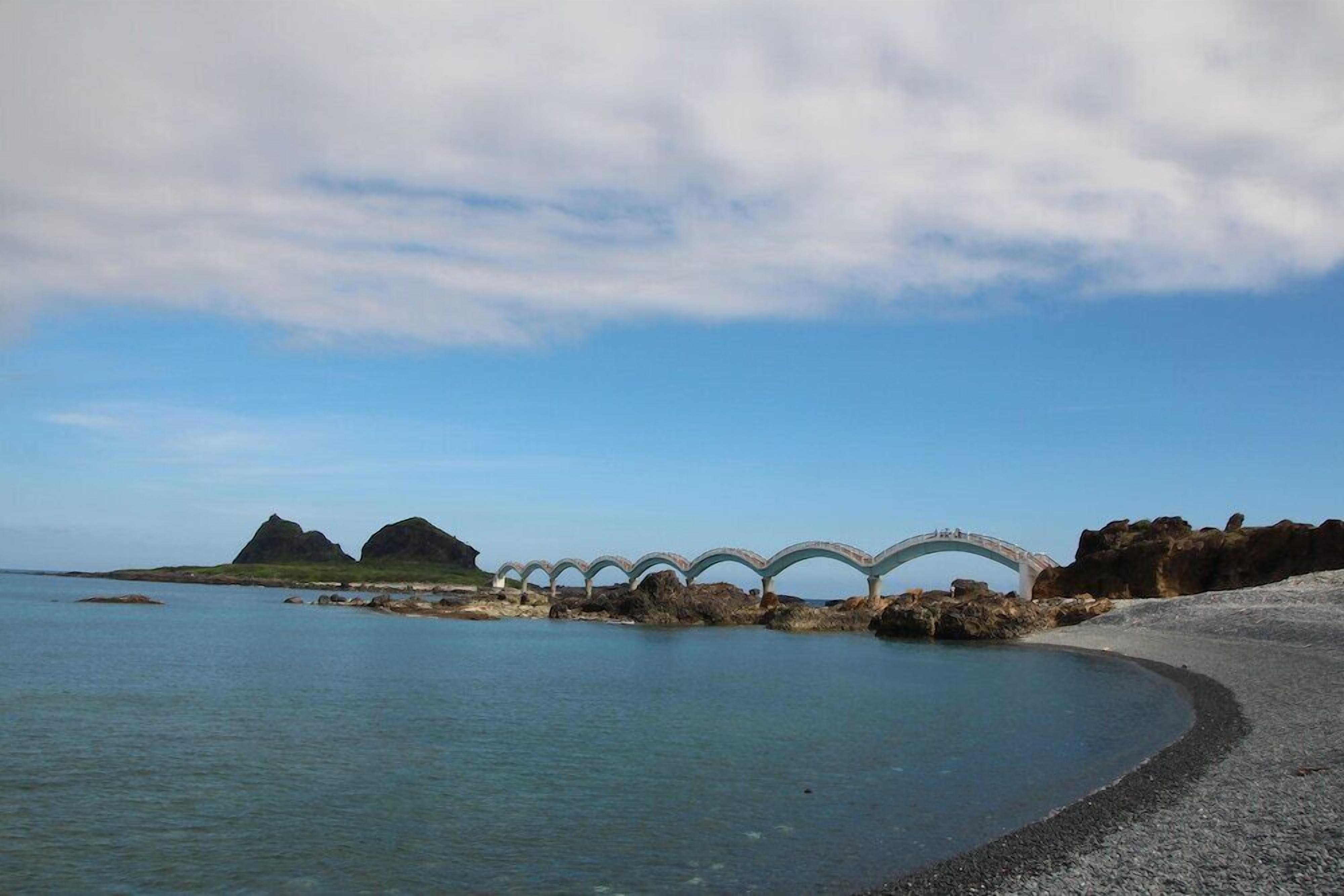 A coastal scene with a white bridge over water, rocky outcrops, and a clear sky.