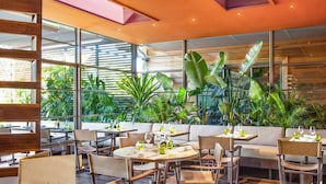 Un restaurante con mesas redondas, sillas y vistas a abundante vegetación a través de amplios ventanales.