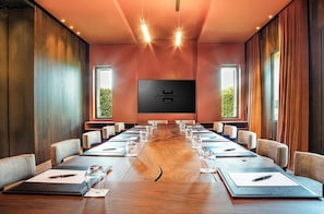 Un salón de conferencias con una larga mesa, sillas, una pantalla grande y ventanas con cortinas.