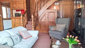 Ein gemütliches Zimmer mit einer Holztreppe, einem Hounds-Tooth-Sessel, einer weißen Couch mit gemusterten Kissen und einem Glastisch mit Tassen und einer Blumenvase.