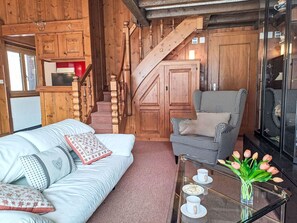Ein gemütliches Zimmer mit einer Holztreppe, einem Hounds-Tooth-Sessel, einer weißen Couch mit gemusterten Kissen und einem Glastisch mit Tassen und einer Blumenvase.
