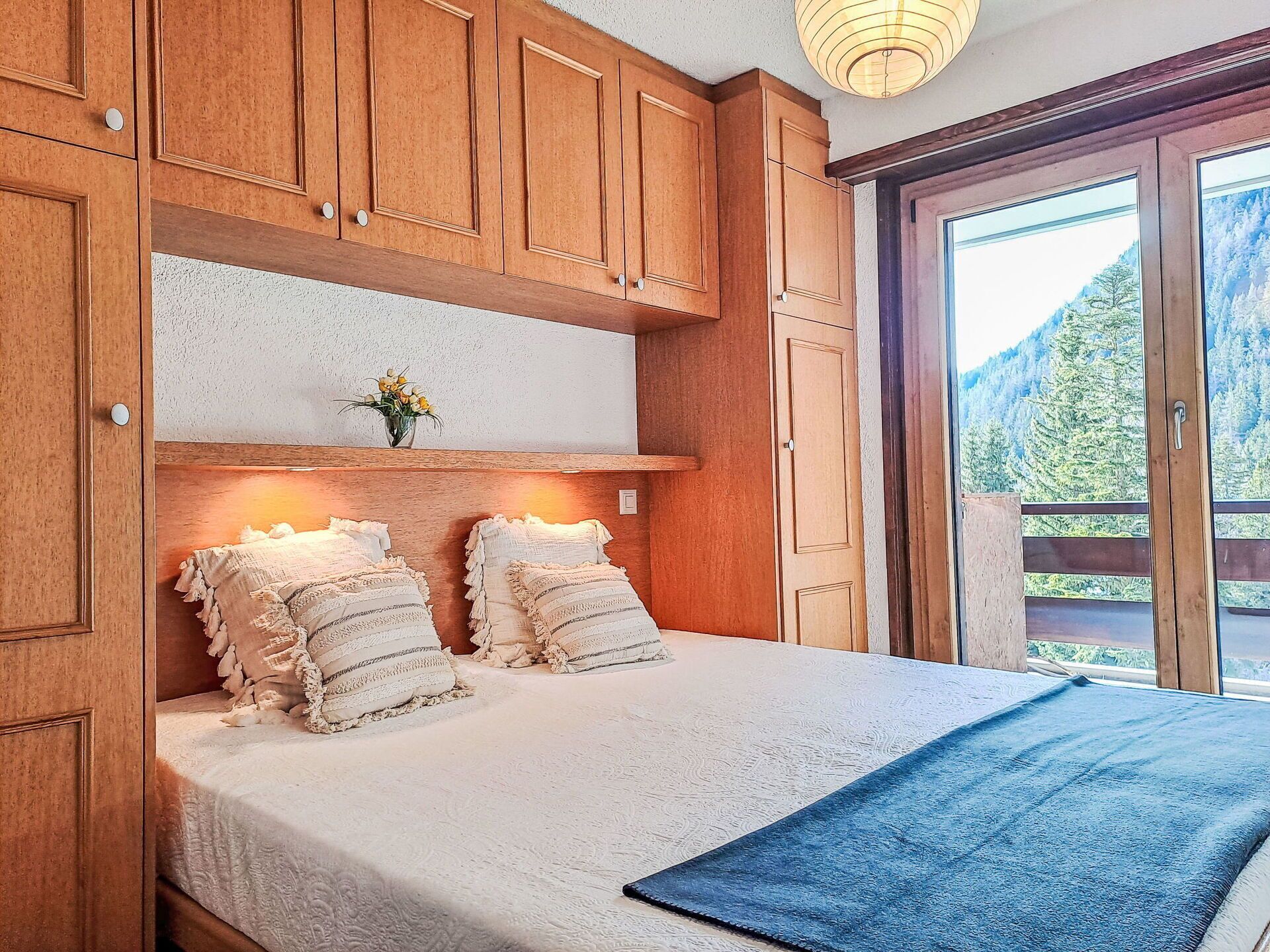 Un dormitorio con armarios de madera, una cama con cubrecama blanco, dos almohadas, un florero con flores y vista a un bosque por una puerta corrediza.