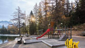 Ein Spielplatz mit einer roten Rutsche, umgeben von Bäumen und einem See, mit Bergen im Hintergrund.