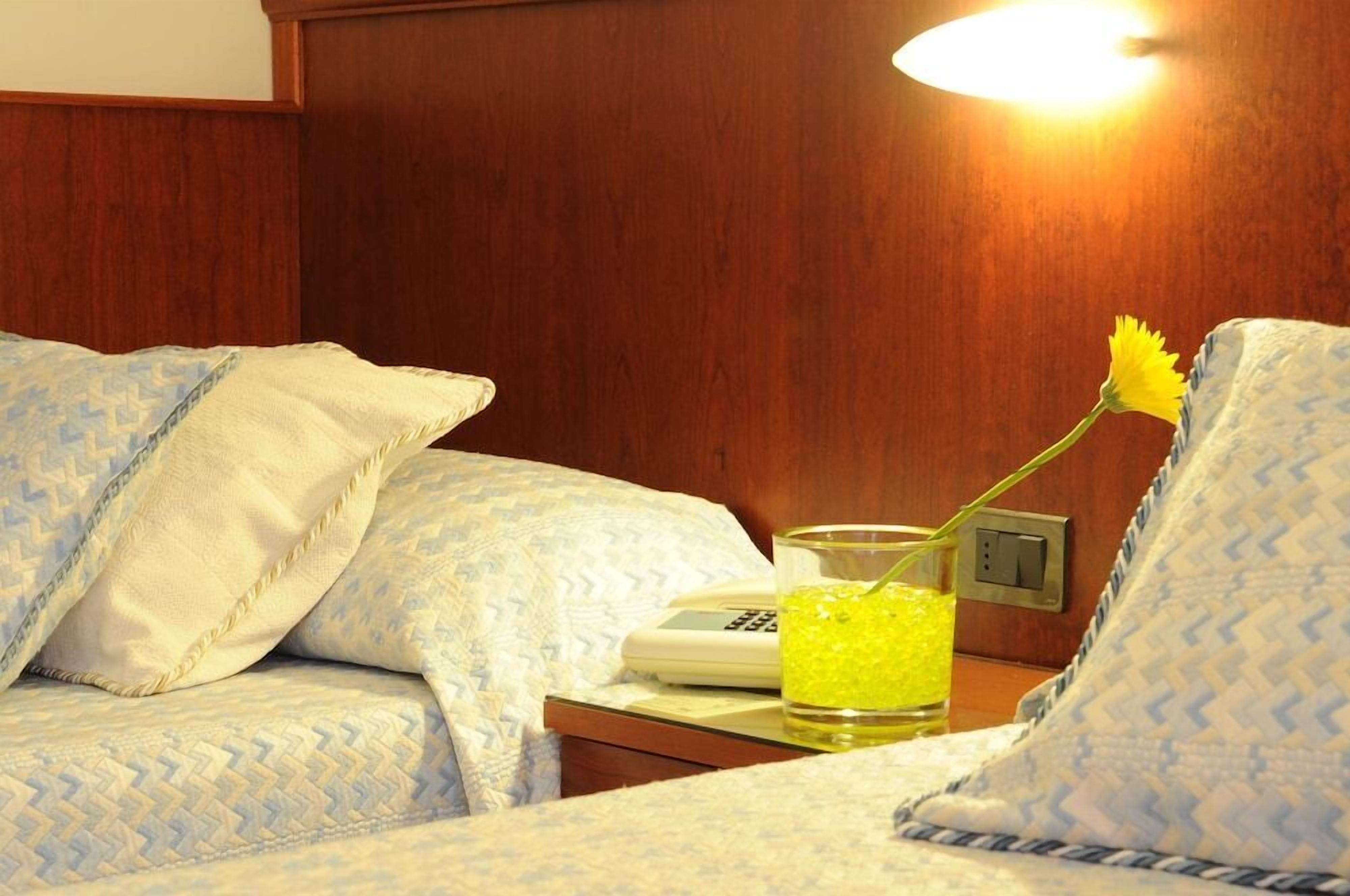 A hotel room with a bed, a bedside table with a telephone and a glass of yellow liquid, and a wall-mounted light.