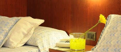 A hotel room with a bed, a bedside table with a telephone and a glass of yellow liquid, and a wall-mounted light.