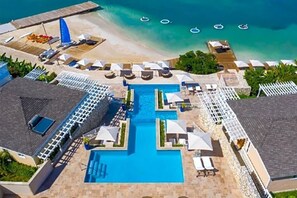 An aerial view of a resort with a central swimming pool, multiple sun loungers, and a beach area with umbrellas.