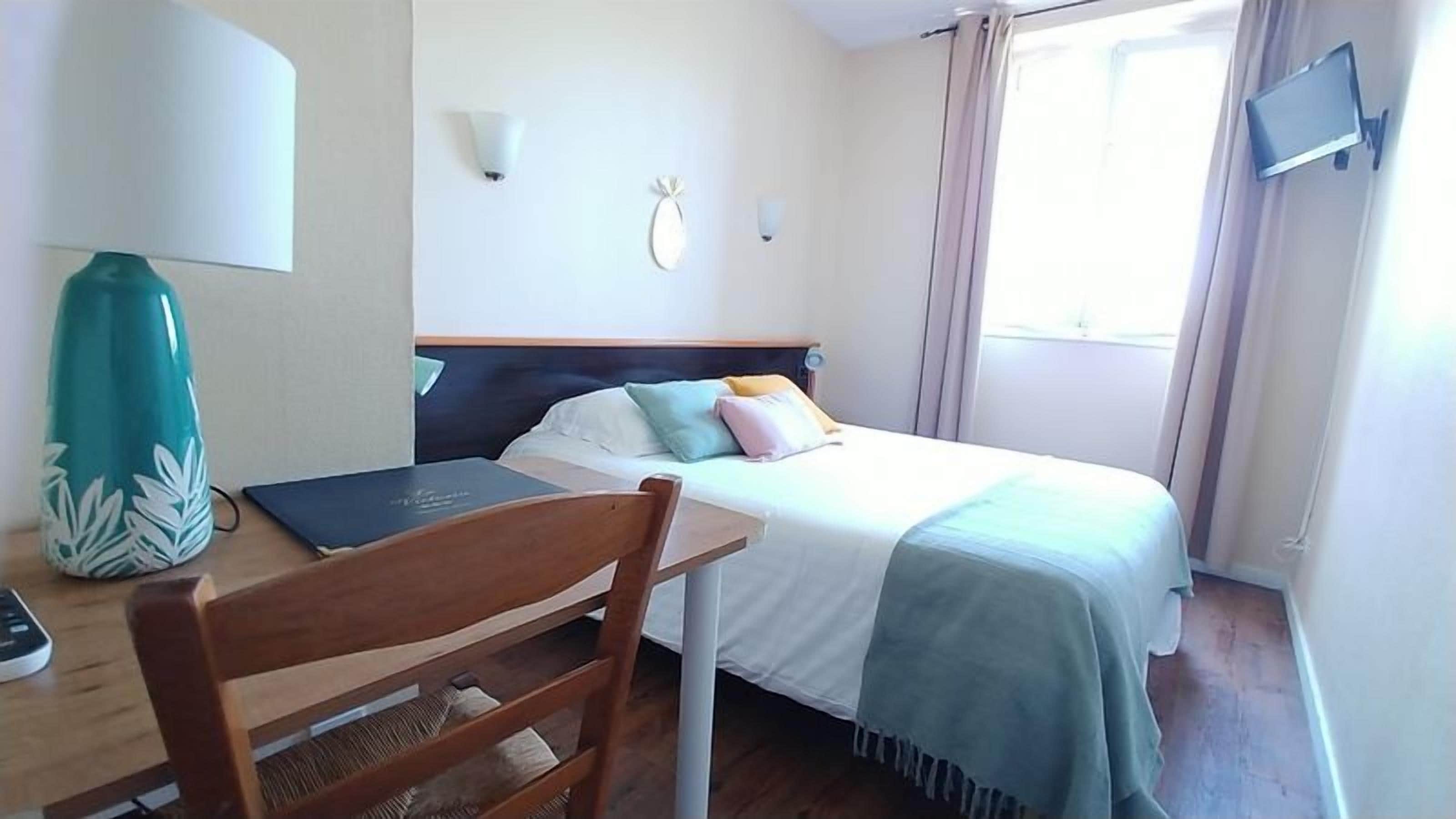 A hotel room with a bed, a desk with a lamp, a chair, a TV mounted on the wall, and a window with curtains.